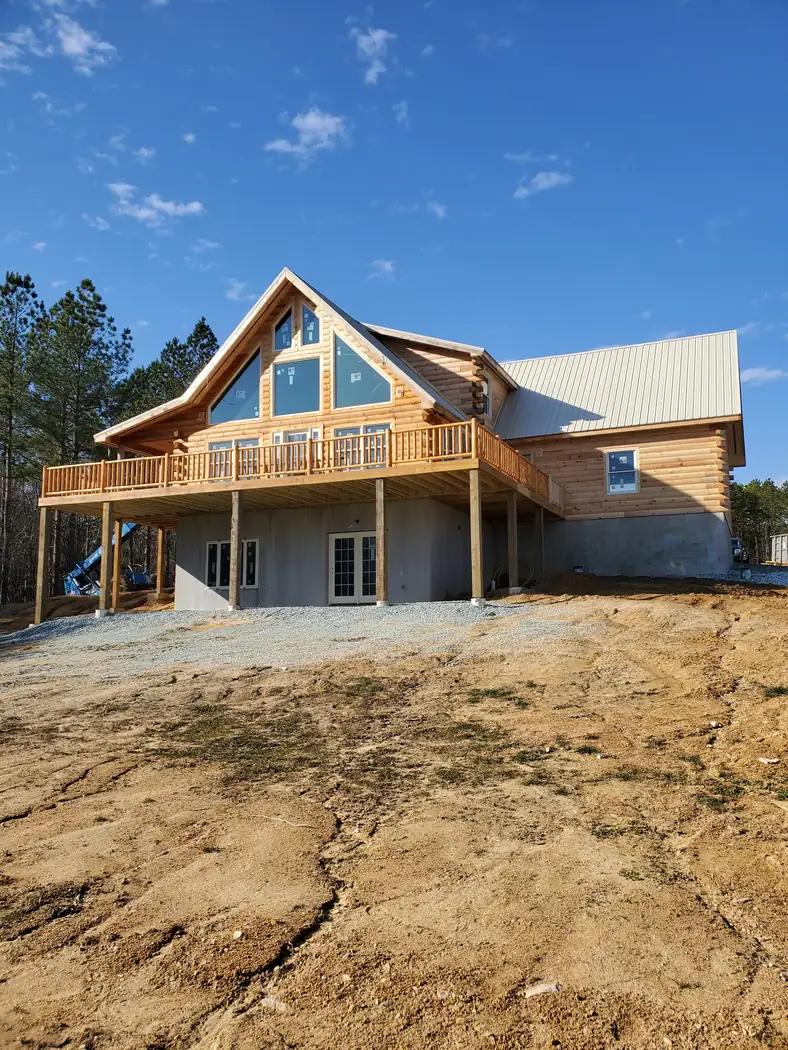 Log home nearing completion with an open elevated deck, tall gable window wall, metal roof, and walkout lower level
