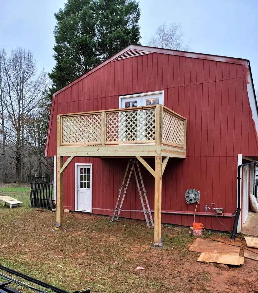 Elevated balcony deck with pressure-treated framing, posts on footings, and lattice railing on a red barn building