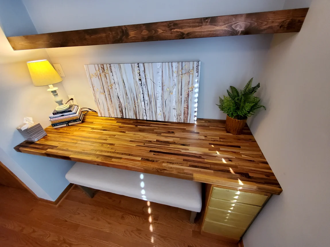 Built-in corner desk with butcher block top beneath stained wood ceiling beams in a blue-walled room