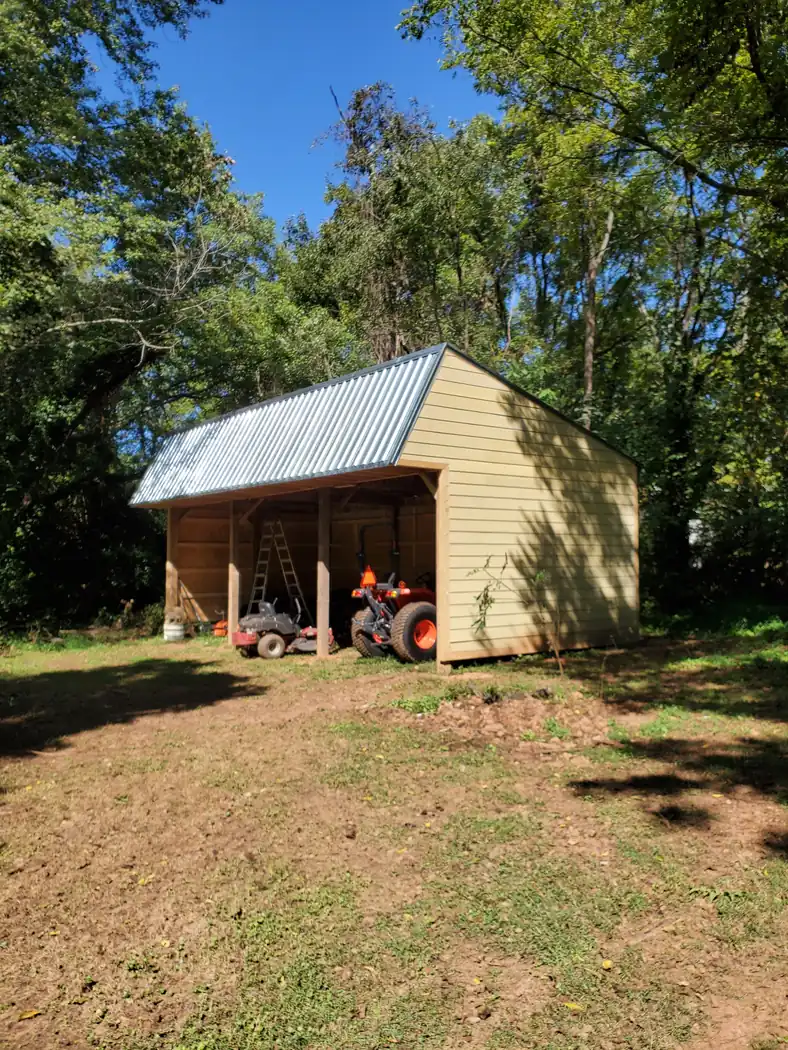 Open-front equipment shed with metal roof and lap siding, sheltering a tractor and riding mower