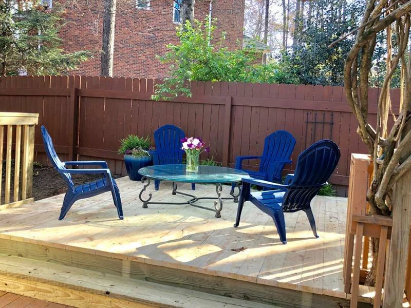 Ground-level wood deck with blue Adirondack chairs and a glass patio table beside a stained privacy fence