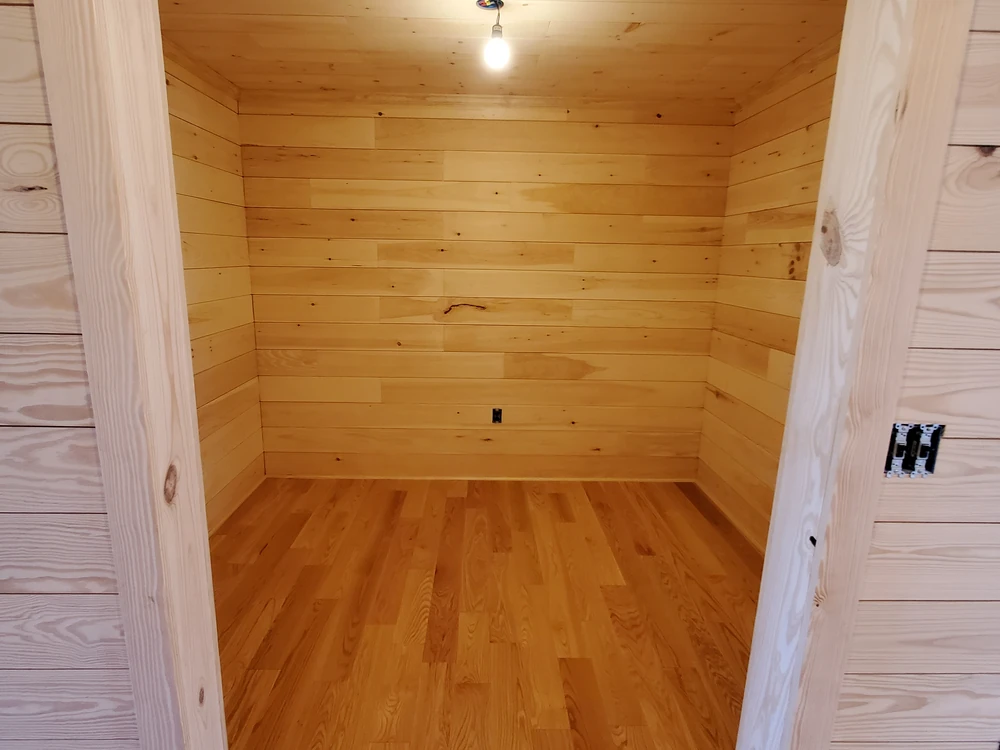 Small empty room clad in knotty wood plank walls and ceiling with new hardwood flooring, trim not yet installed