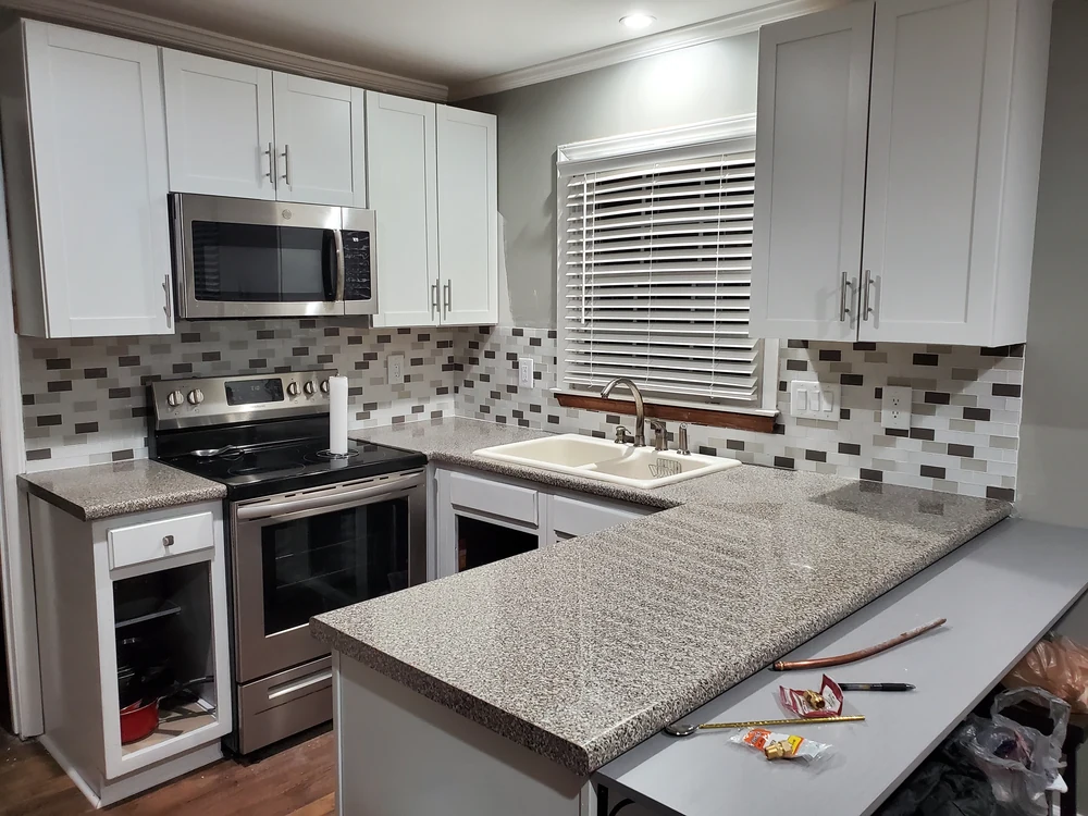 White shaker kitchen with speckled stone-look counters and glass mosaic backsplash mid-installation