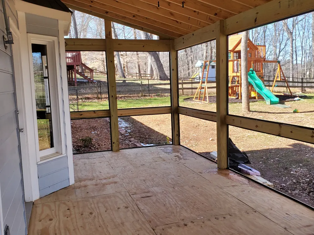 Screened porch interior mid-build with bare plywood subfloor, exposed framing, and screens