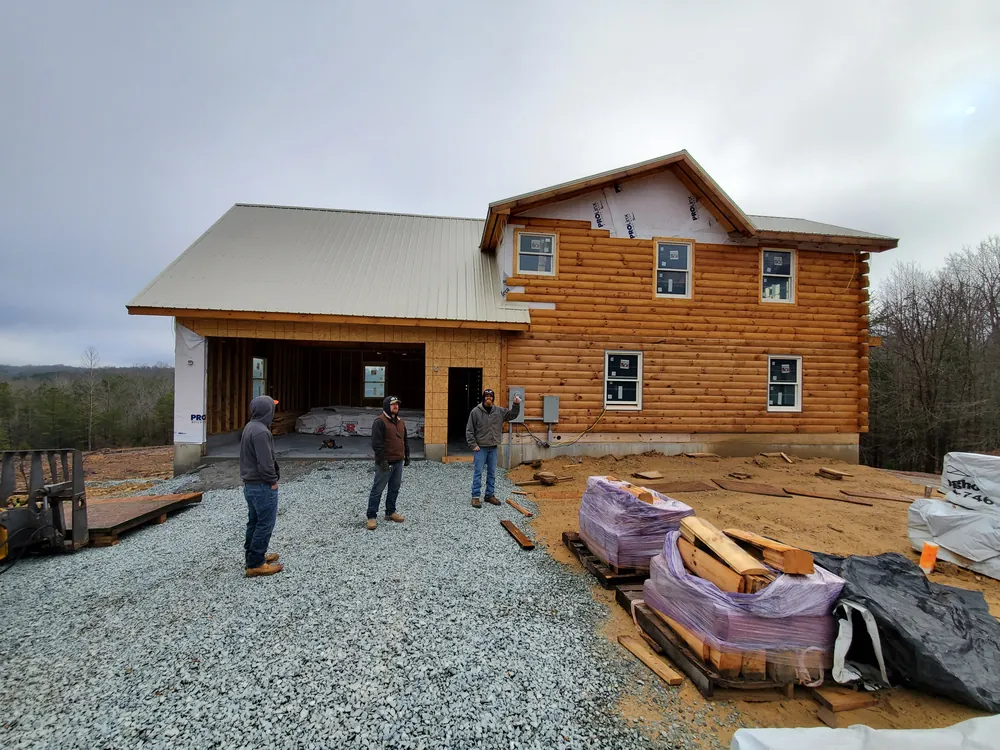 Two-story log-sided home under construction with a metal roof, open garage bays, and workers on a gravel drive