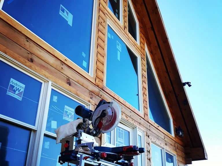 New wood siding courses and window trim going up over blue housewrap around new windows, miter saw on site